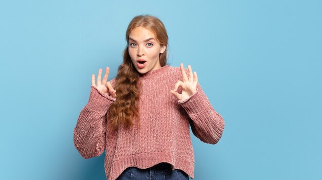 Mujer bonita rubia que se siente conmocionada, asombrada y sorprendida, mostrando aprobación haciendo un signo de bien con ambas manos