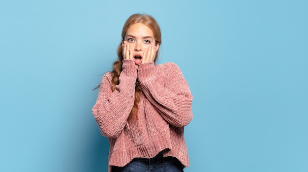 Mujer bonita rubia que se siente conmocionada y asustada, mirando aterrorizada con la boca abierta y las manos en las mejillas