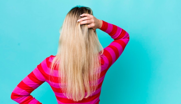 Mujer bonita rubia que se siente despistada y confundida pensando en una solución con la mano en la cadera y otra en la vista trasera de la cabeza