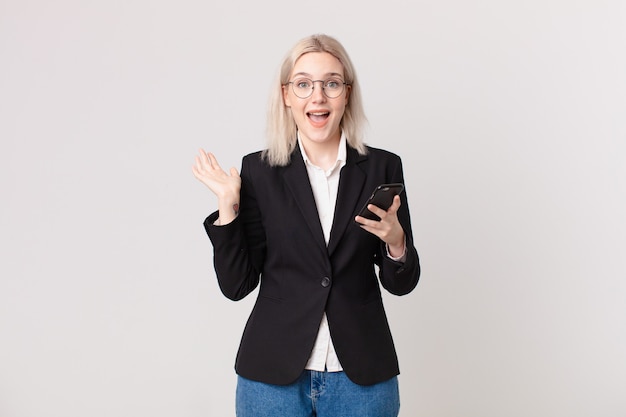 Mujer bonita rubia que se siente feliz y asombrada por algo increíble y sostiene un teléfono móvil