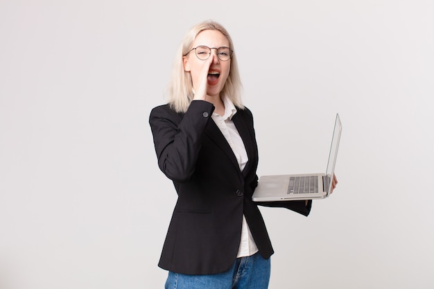 Mujer bonita rubia que se siente feliz, dando un gran grito con las manos al lado de la boca y sosteniendo una computadora portátil