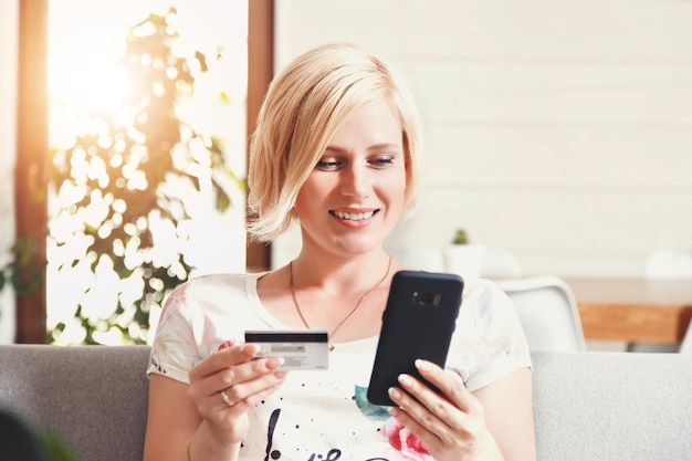 Mujer bonita rubia sentada en el sofá y haciendo compras con el teléfono inteligente usando el concepto de compra en línea con tarjeta de crédito