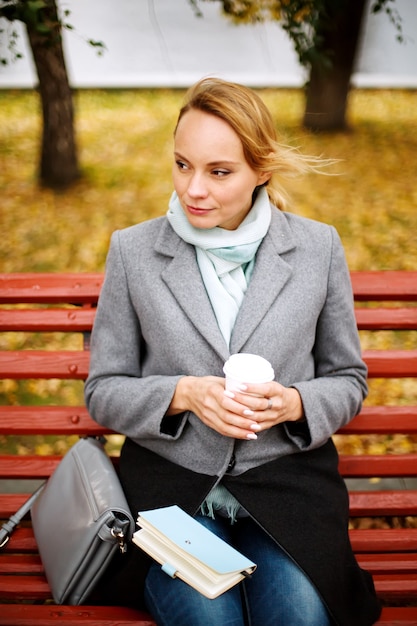 Mujer bonita rubia sentada sola en un banco en el parque de otoño con un cuaderno y una taza de bebida