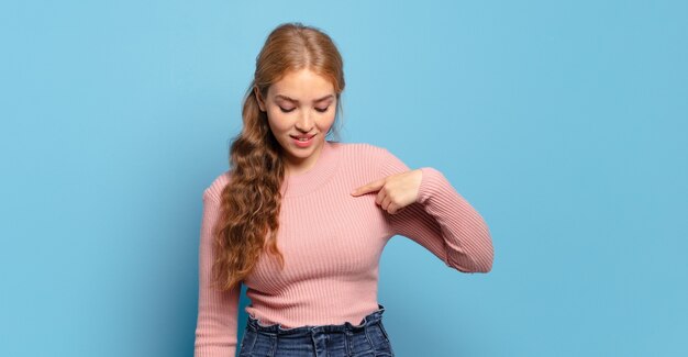 Mujer bonita rubia sonriendo alegre y casualmente, mirando hacia abajo y apuntando al pecho