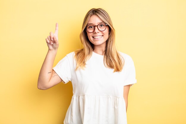 Mujer bonita rubia sonriendo alegre y felizmente, apuntando hacia arriba con una mano para copiar el espacio