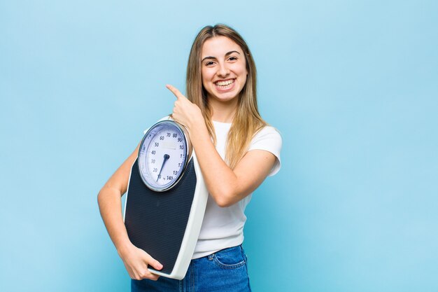 Mujer bonita rubia sonriendo alegremente, sintiéndose feliz y apuntando hacia un lado y hacia arriba, mostrando el objeto en el espacio de la copia