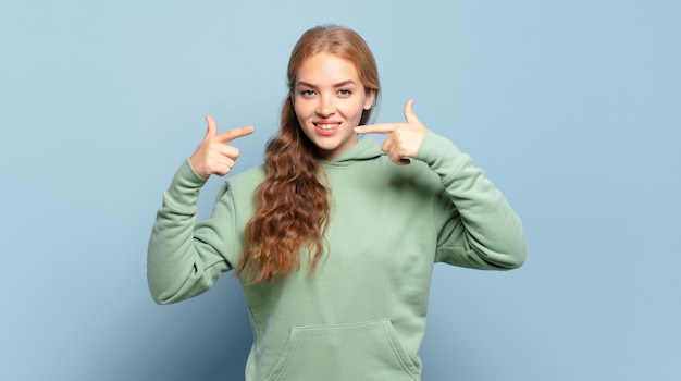 Mujer bonita rubia sonriendo con confianza apuntando a su propia sonrisa amplia, actitud positiva, relajada y satisfecha