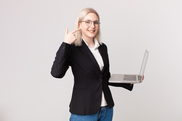 Mujer bonita rubia sonriendo con confianza apuntando a su propia sonrisa amplia y sosteniendo una computadora portátil