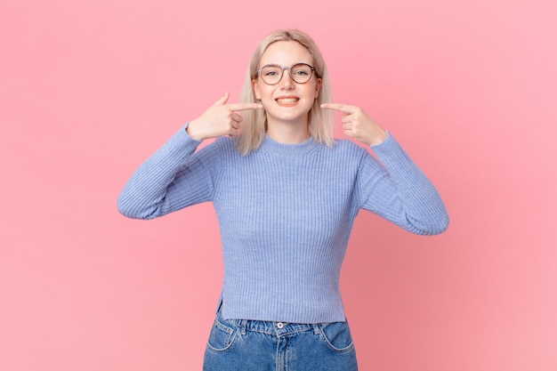 Mujer bonita rubia sonriendo con confianza apuntando a su propia sonrisa amplia