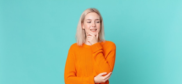 Mujer bonita rubia sonriendo con una expresión feliz y segura con la mano en la barbilla