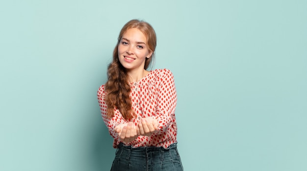 Mujer bonita rubia sonriendo felizmente con mirada amigable, segura y positiva, ofreciendo y mostrando un objeto o concepto