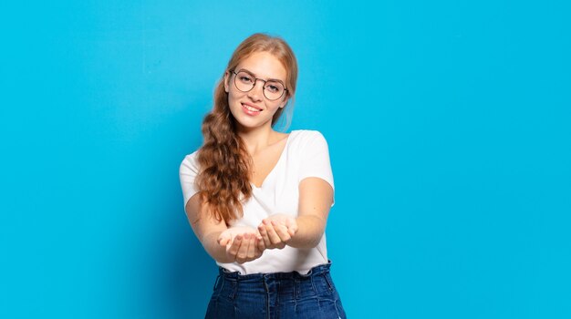 Mujer bonita rubia sonriendo felizmente con mirada amistosa, segura y positiva, ofreciendo y mostrando un objeto o concepto