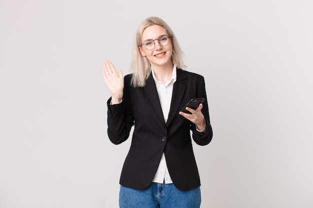 Mujer bonita rubia sonriendo felizmente, saludando con la mano, dándote la bienvenida y saludándote y sosteniendo un teléfono móvil