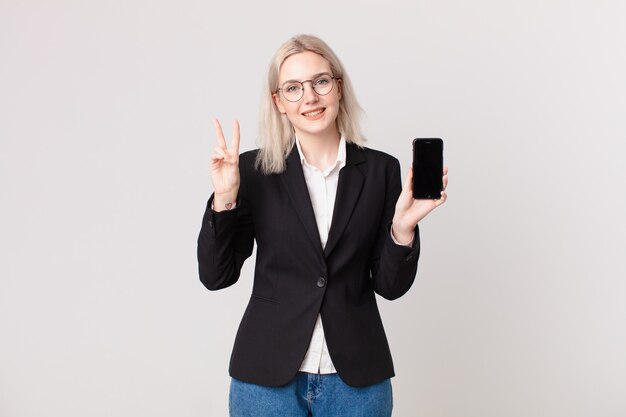 Mujer bonita rubia sonriendo y mirando amigable, mostrando el número dos y sosteniendo un teléfono móvil