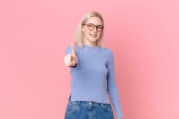 Mujer bonita rubia sonriendo con orgullo y confianza haciendo el número uno