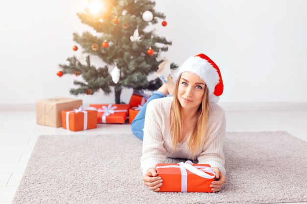 Mujer bonita rubia con un suéter de punto cálido, acostado sobre una alfombra cerca del árbol de Navidad decorado en una sala de estar. Muchos regalos debajo del árbol.