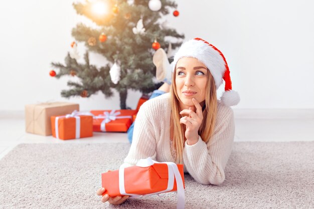 Mujer bonita rubia con un suéter de punto cálido, acostado sobre una alfombra cerca del árbol de Navidad decorado en una sala de estar. Muchos regalos debajo del árbol.