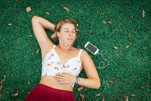 Mujer bonita rubia tumbada en el césped con los ojos cerrados escuchando música con teléfono móvil