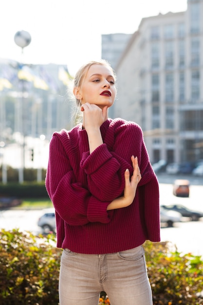 Mujer bonita rubia vestida con jeans grises y suéter de punto, posando en la ciudad