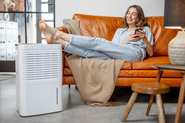 Mujer bonita sentada cerca del aparato purificador de aire y humectante cerca del sofá que monitorea la calidad del aire en el teléfono. Microclima de salud en el concepto de hogar.