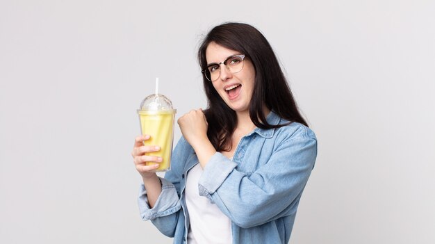 Mujer bonita sintiéndose feliz y enfrentando un desafío o celebrando y sosteniendo un batido de vainilla