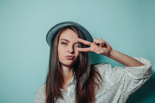 Mujer bonita en un sombrero gris con cabello largo y camisa blanca. La mujer muestra el signo de la paz con los dedos.