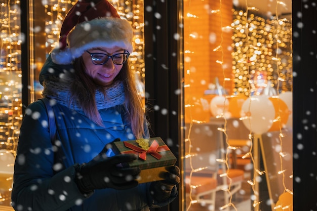 Mujer bonita con sombrero de Papá Noel con caja de regalos cerca de la ventana del café iluminado