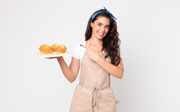 Mujer bonita sonriendo alegremente, sintiéndose feliz y apuntando hacia un lado y sosteniendo una bandeja de muffins