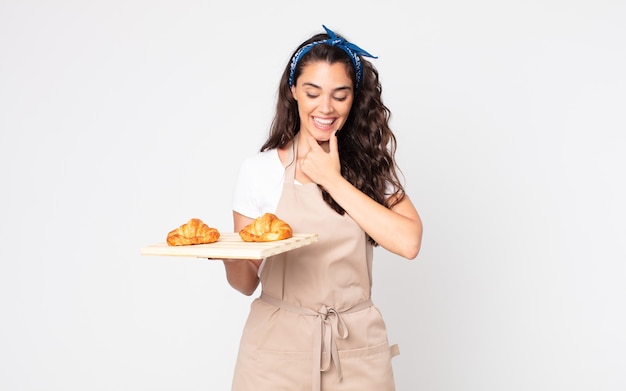 Mujer bonita sonriendo con una expresión feliz y segura con la mano en la barbilla y sosteniendo una bandeja de croissants