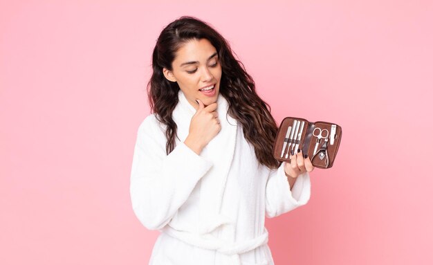 Mujer bonita sonriendo con una expresión feliz y segura con la mano en la barbilla y sosteniendo una bolsa de maquillaje con herramientas de uñas