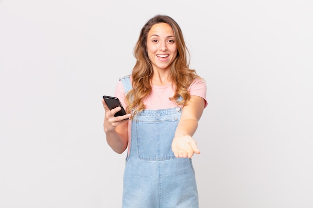 Mujer bonita sonriendo felizmente con amigable y ofreciendo y mostrando un concepto y sosteniendo un teléfono inteligente