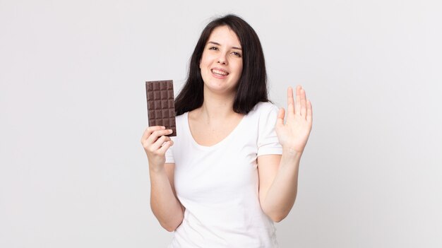 Mujer bonita sonriendo felizmente, saludando con la mano, dándote la bienvenida y saludándote y sosteniendo una barra de chocolate