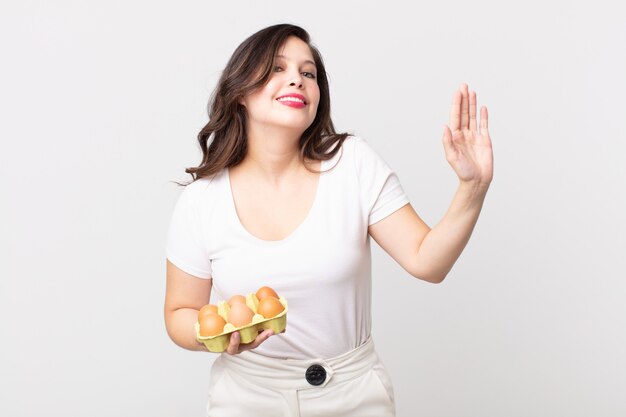 Mujer bonita sonriendo felizmente, saludando con la mano, dándote la bienvenida y saludándote y sosteniendo una caja de huevos