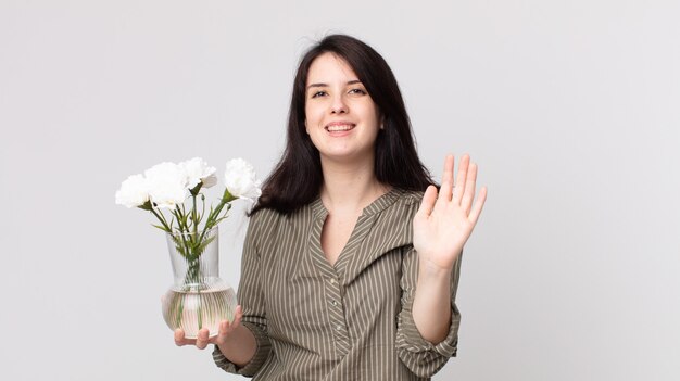 Mujer bonita sonriendo felizmente, saludando con la mano, dándote la bienvenida y saludándote y sosteniendo flores decorativas. agente asistente con un auricular