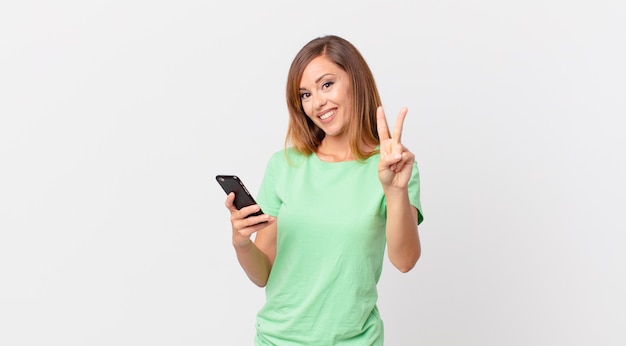 Mujer bonita sonriendo y mirando amigable, mostrando el número dos y usando un teléfono inteligente