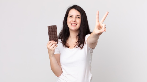 Mujer bonita sonriendo y mirando feliz, gesticulando victoria o paz y sosteniendo una barra de chocolate