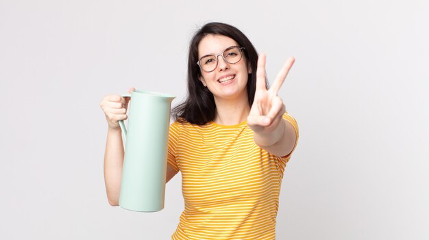 Mujer bonita sonriendo y mirando feliz, gesticulando victoria o paz y sosteniendo un termo de café