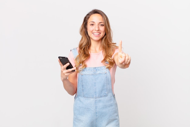 Mujer bonita sonriendo con orgullo y confianza haciendo el número uno y sosteniendo un teléfono inteligente