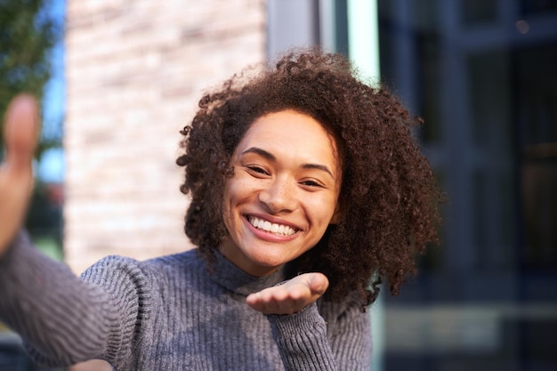 Mujer bonita sonriendo con una sonrisa con dientes que sopla un beso de aire en una cámara imaginaria en su mano extendida