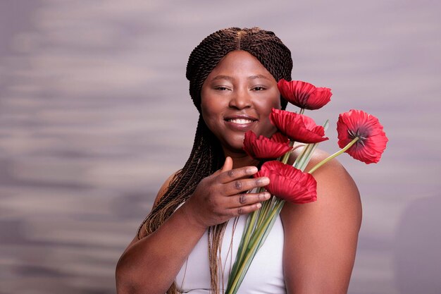 Mujer bonita sonriente posando con un retrato de rama de amapola roja. Joven modelo afroamericano de talla grande sosteniendo un símbolo fiminino, de pie con una flor y mirando la cámara en el estudio