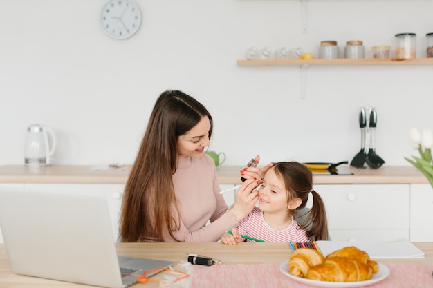 La mujer bonita sonriente que hace compone mientras que se sienta con su pequeña hija en la cocina durante el desayuno.