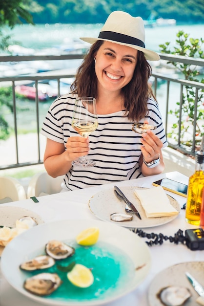 Mujer bonita sonriente con sombrero fedora sentada en un restaurante al aire libre comiendo ostras y bebiendo vino