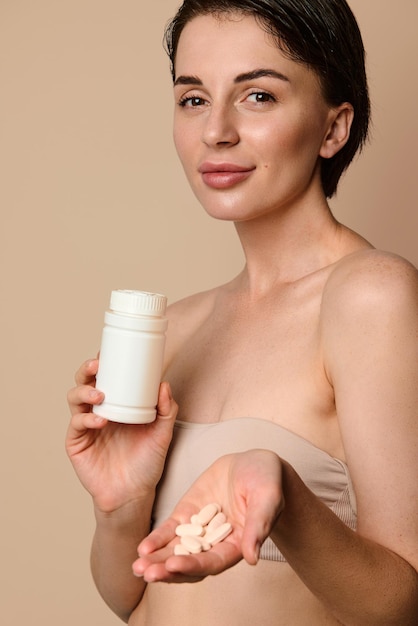 Mujer bonita sosteniendo una botella con suplemento dietético, vitaminas y minerales y mostrando algunas pastillas en sus manos, sonriendo tiernamente mirando a la cámara. Concepto de cuidado de la salud, la piel y el cuerpo.