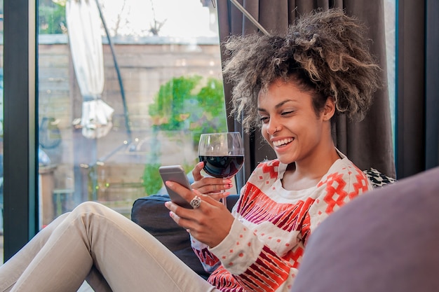 Mujer bonita en su sala de estar acostado en el sofá enviando messages.Portrait de una mujer africana mirando teléfono móvil y disfrutar de vaso de vino