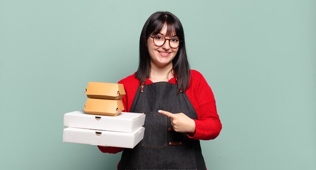 Mujer bonita de talla grande sonriendo alegremente, sintiéndose feliz y apuntando hacia un lado y hacia arriba, mostrando el objeto en el espacio de la copia