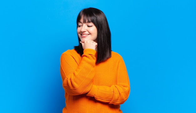 Mujer bonita de talla grande sonriendo con una expresión feliz y segura con la mano en la barbilla, preguntándose y mirando hacia un lado