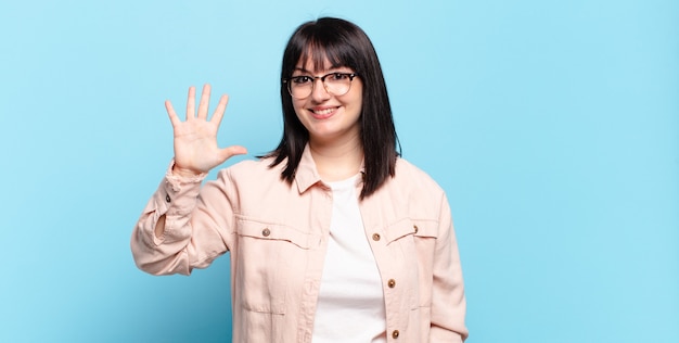 Mujer bonita de talla grande sonriendo y mirando amigable, mostrando el número cinco o quinto con la mano hacia adelante, contando hacia atrás