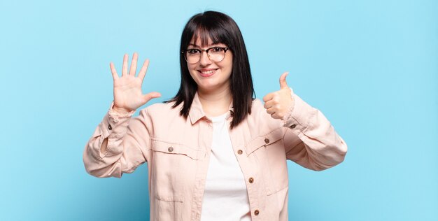 Mujer bonita de talla grande sonriendo y mirando amigable, mostrando el número seis o sexto con la mano hacia adelante, contando hacia atrás