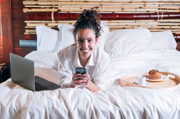 Mujer bonita con teléfono y computadora portátil en la cama