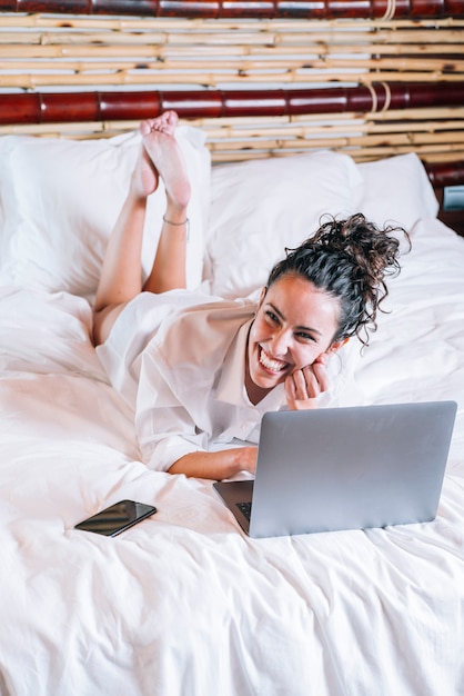 Mujer bonita con teléfono y computadora portátil en la cama